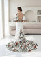 White crepe mermaid wedding dress with open back, flutter sleeves, and colorful earthy floral embroidery on skirt.