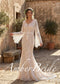 Boho ivory lace mermaid wedding dress with V-neck, tassel tie, and long bell sleeves, worn in a rustic stone courtyard.