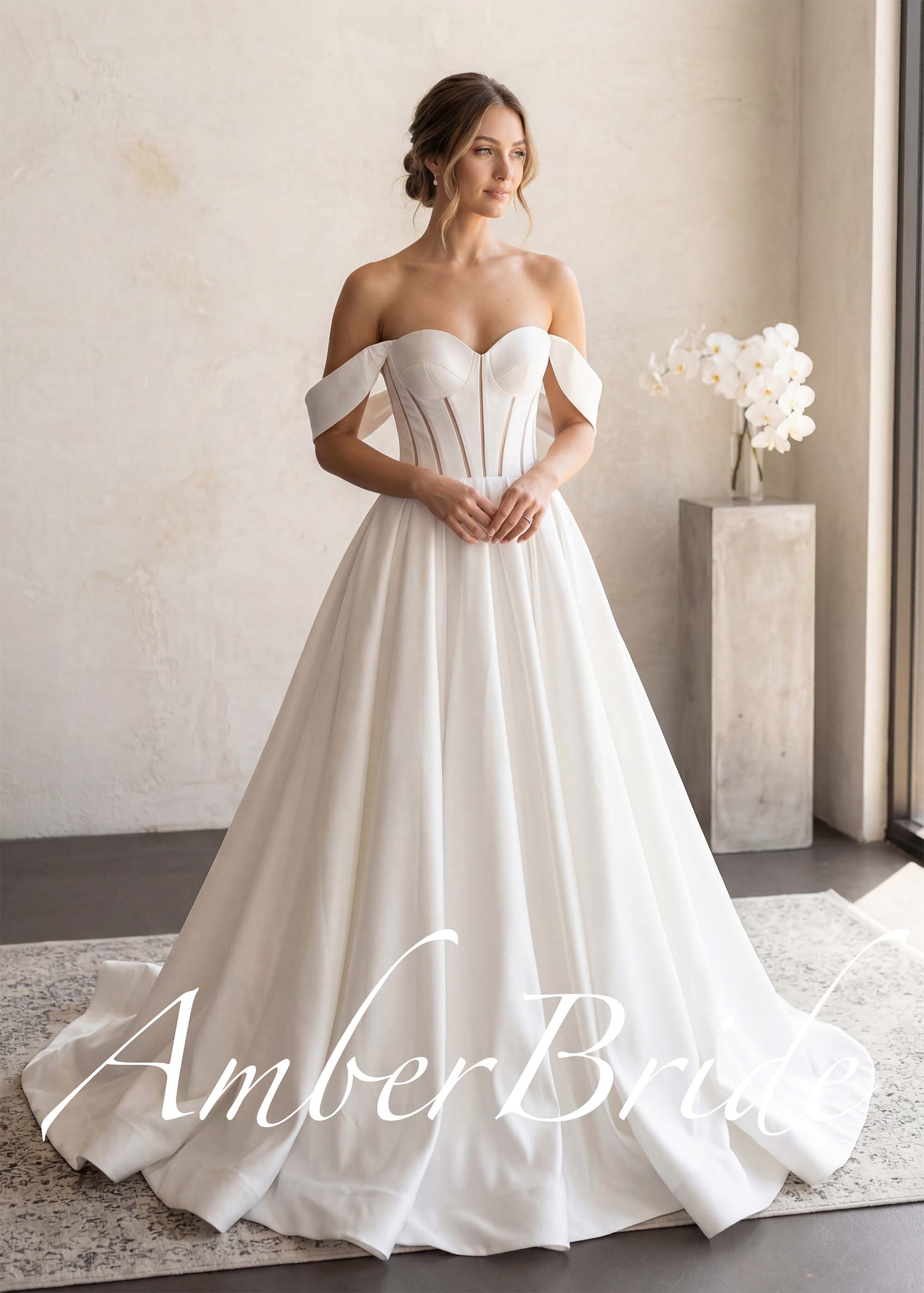 White satin A-line wedding dress with off-shoulder sleeves and a structured, semi-sheer boned bodice.