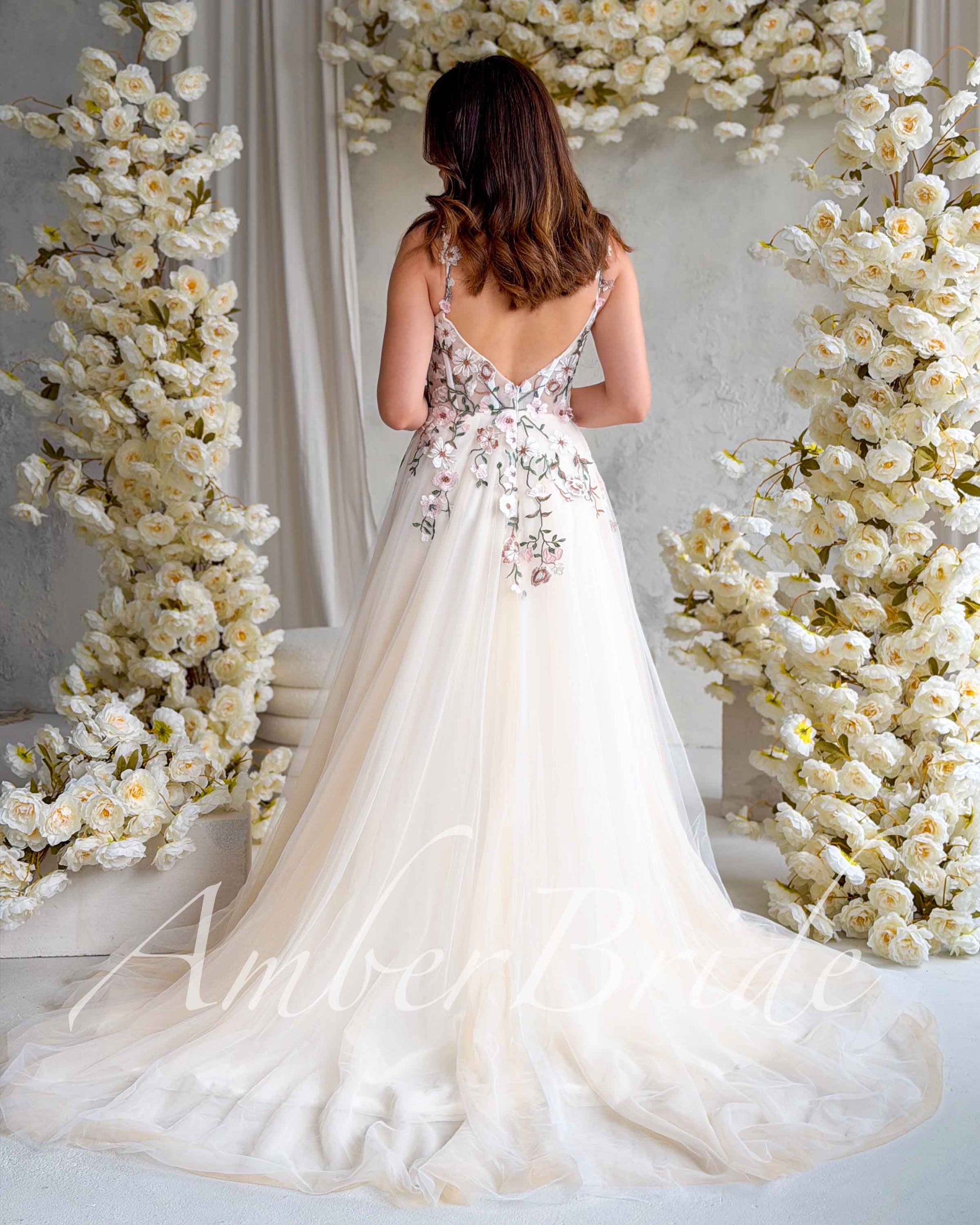 Nude A-line wedding dress with open back, floral pastel embroidery, and flowing tulle train between flower arches.