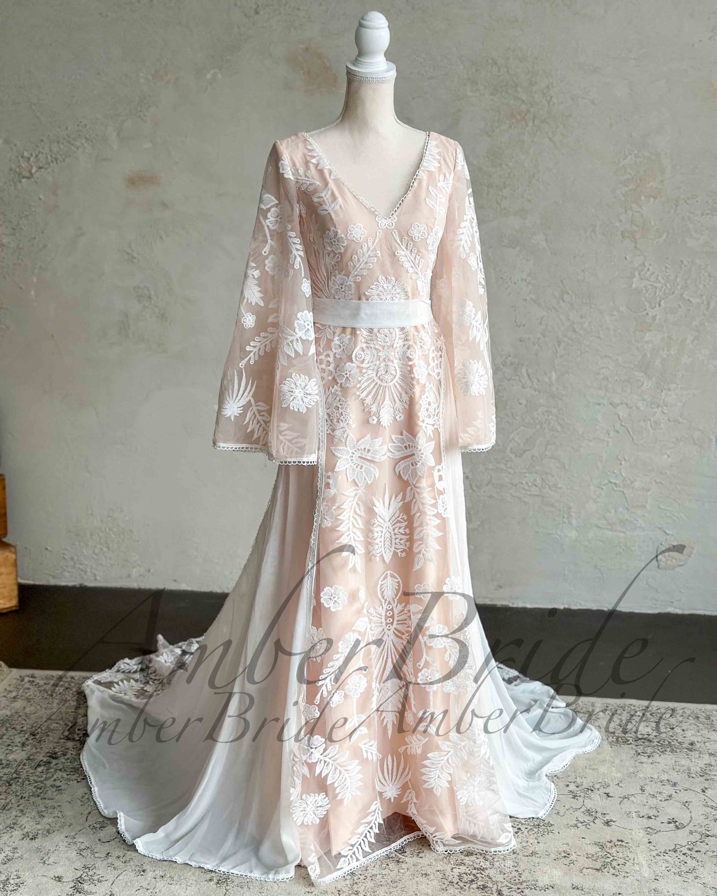 Nude boho wedding dress with white floral embroidery, V-neckline, long cape sleeves, and a defined waistline.