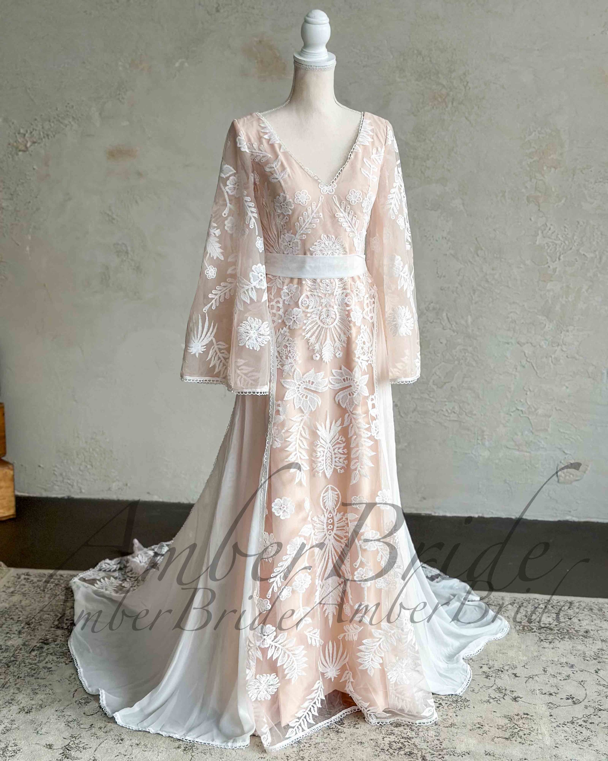 Nude boho wedding dress with white floral embroidery, V-neckline, long cape sleeves, and a defined waistline.