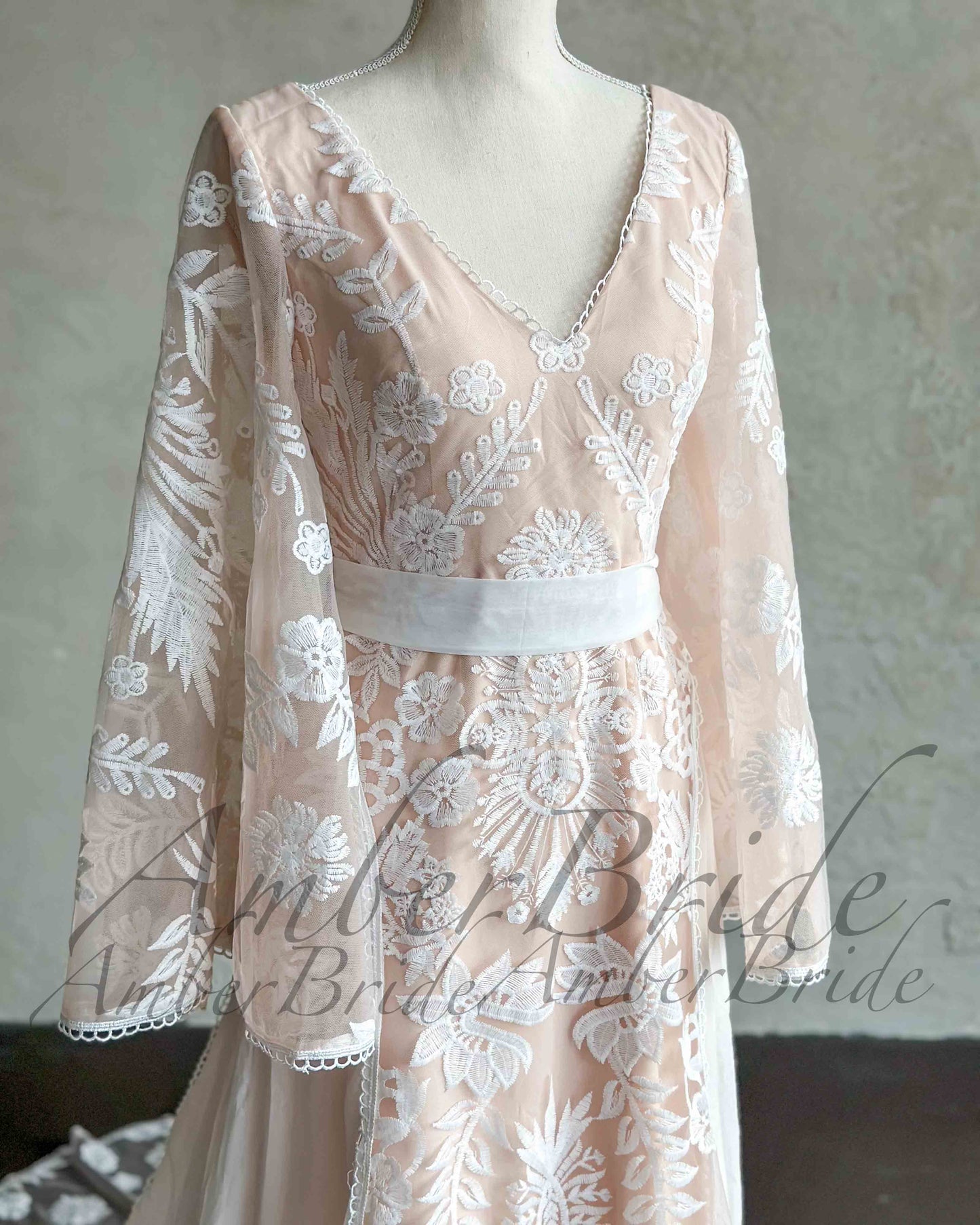 Nude boho wedding dress with white floral embroidery, V-neckline, cape sleeves, and a white satin waist belt.