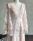 Nude boho wedding dress with white floral embroidery, V-neckline, cape sleeves, and a white satin waist belt.