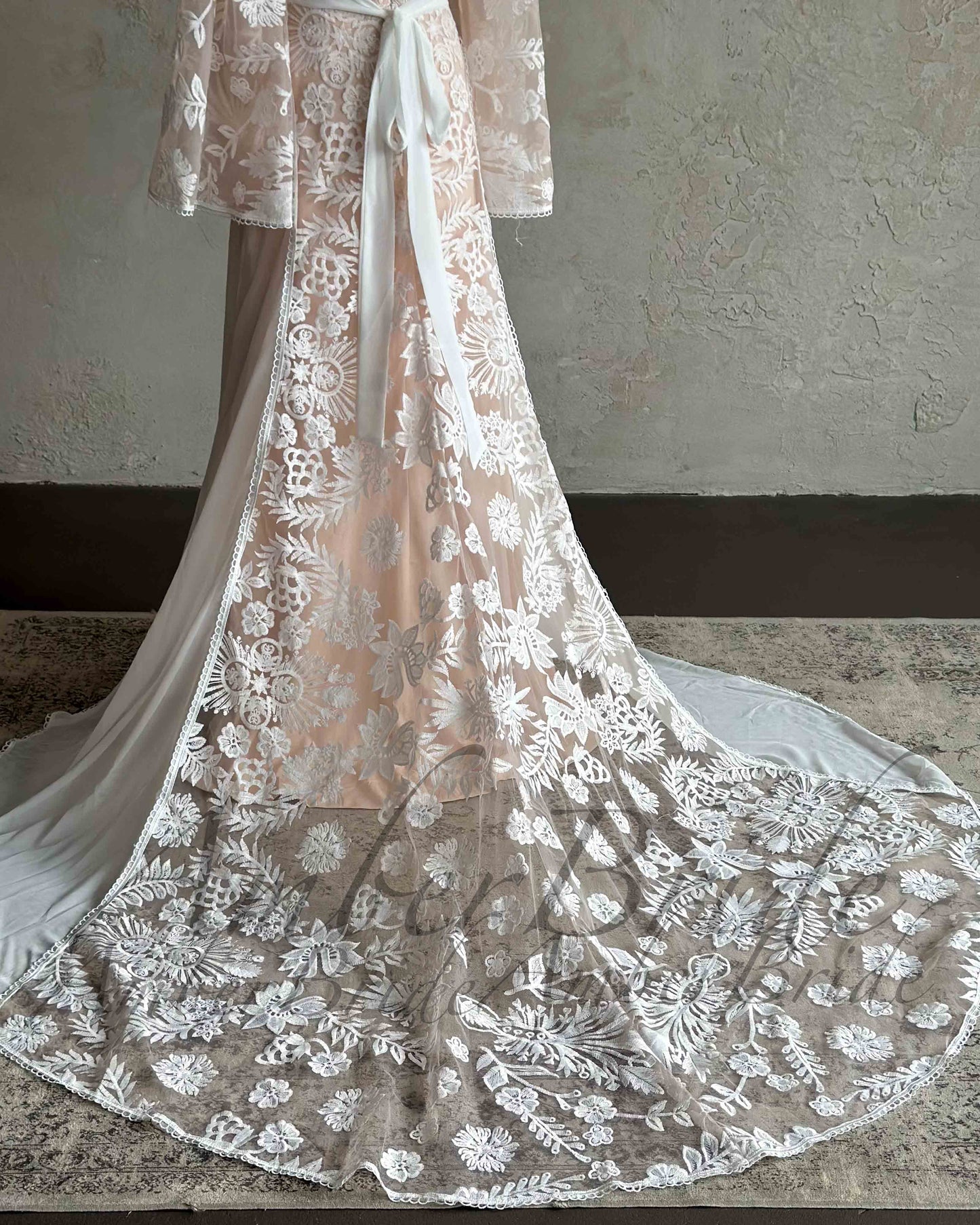 Boho wedding dress with a peach-toned slip, white floral embroidery, cape sleeves, and a long, flowing lace train.