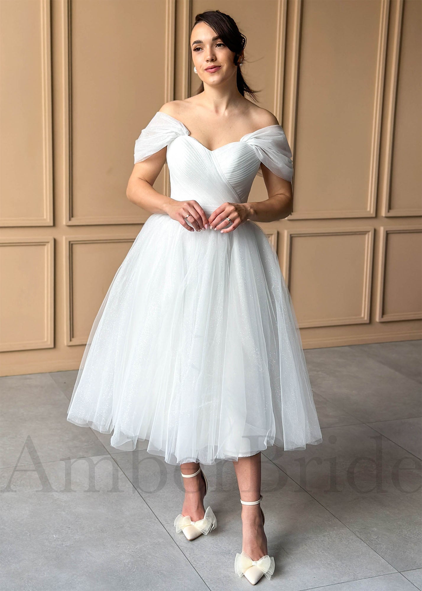 White tea-length glitter tulle wedding dress with off-shoulder sleeves, sweetheart corset bodice, and full skirt.
