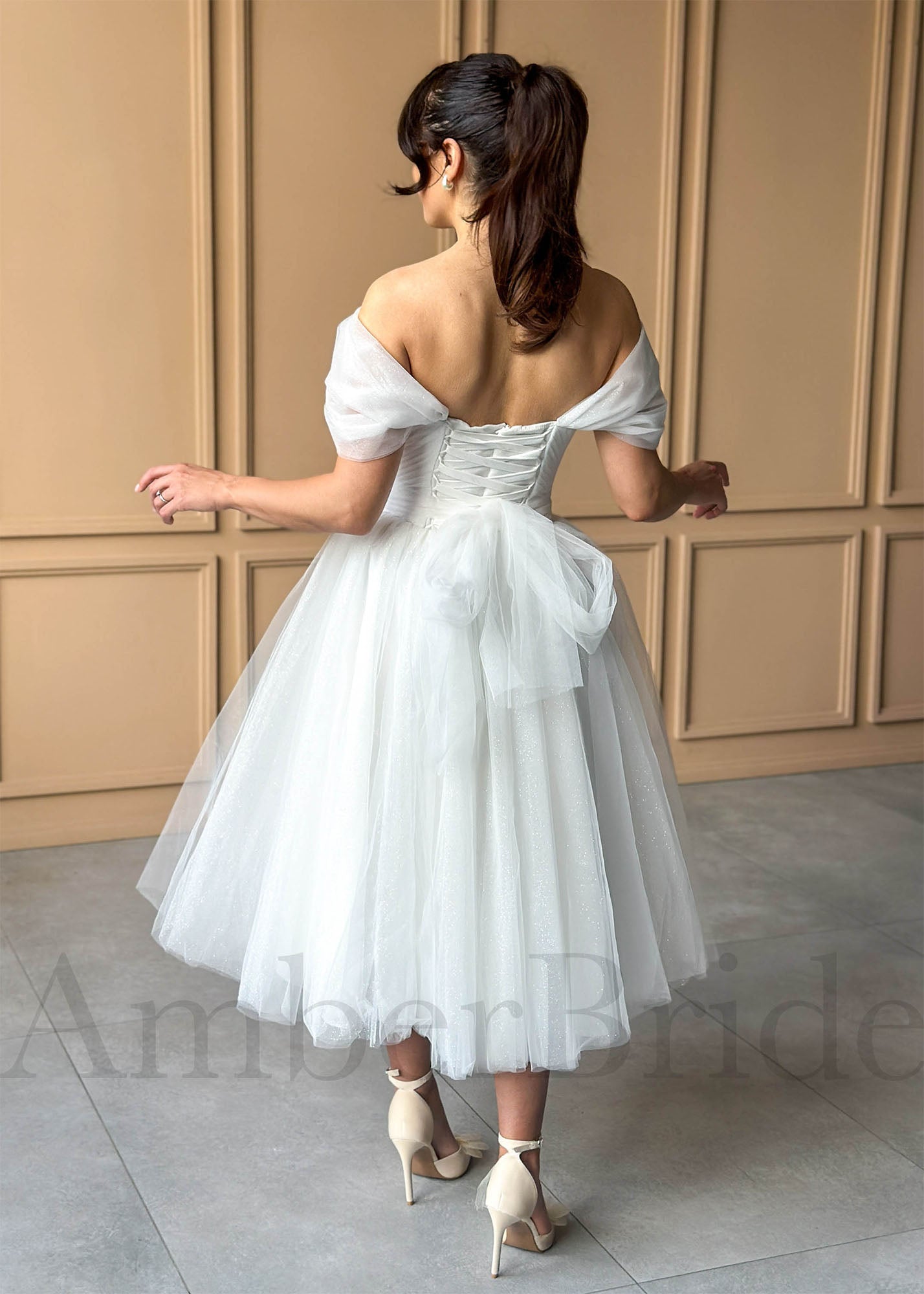 White tea-length glitter tulle dress with off-shoulder sleeves, corset back, and a tulle bow at the waist.