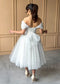 White tea-length glitter tulle dress with off-shoulder sleeves, corset back, and a tulle bow at the waist.