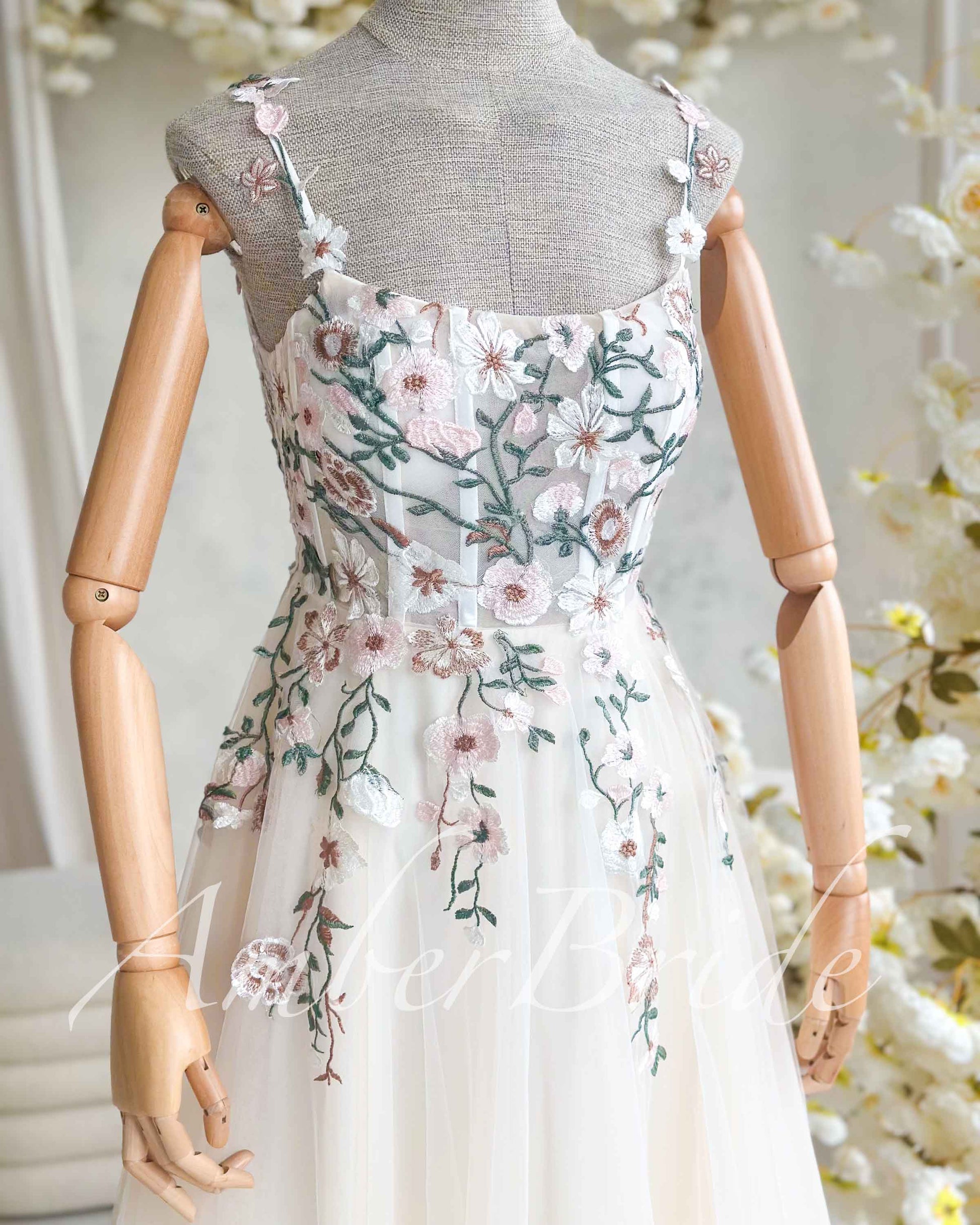 Nude wedding dress on a mannequin with square neckline, spaghetti straps, and pastel floral embroidery on tulle.