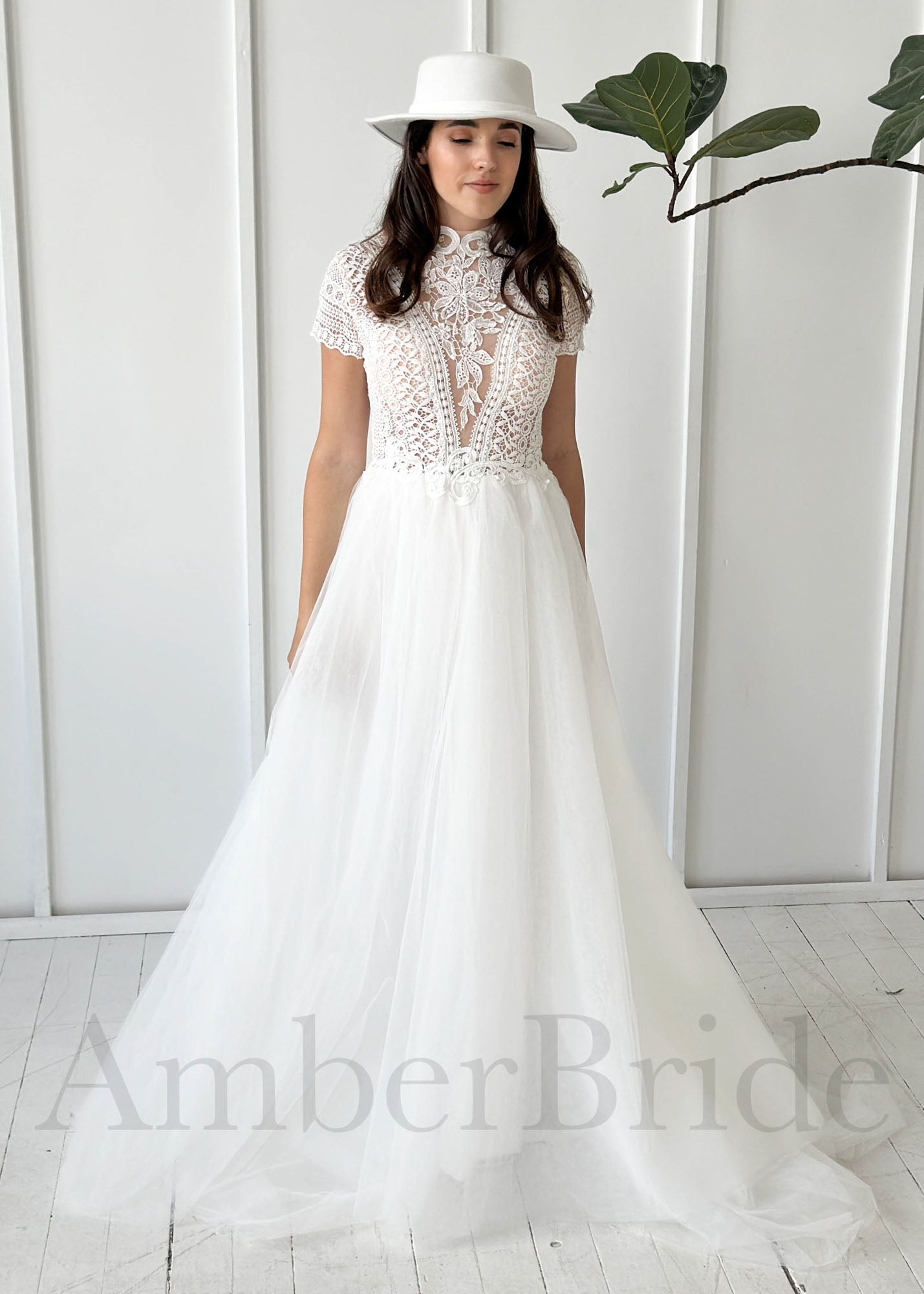 A-line wedding dress with a high-neck lace bodice, short sleeves, and a full tulle skirt, worn with a white hat.